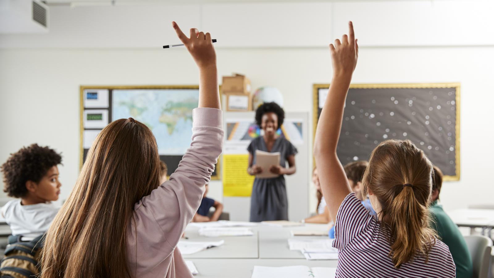 Two students in a high school classroom, with raised hands and a teacher smiling in background